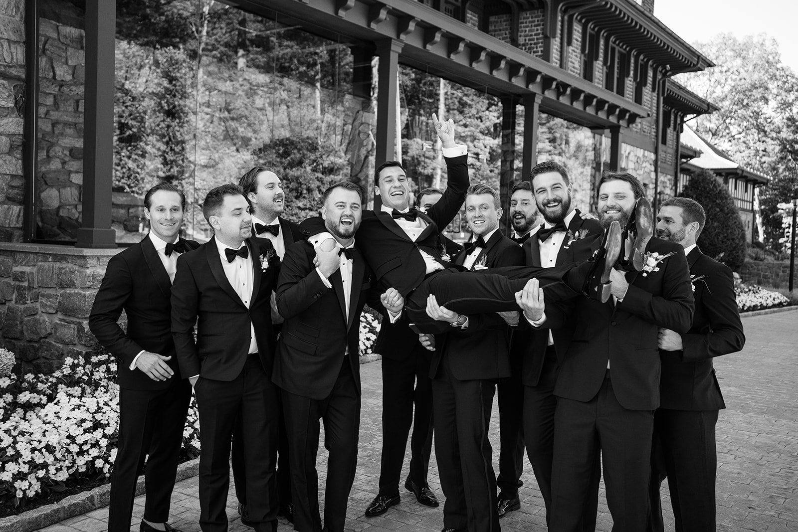 Black and white film photo of the groomsmen playfully lifting the laughing groom into the air.